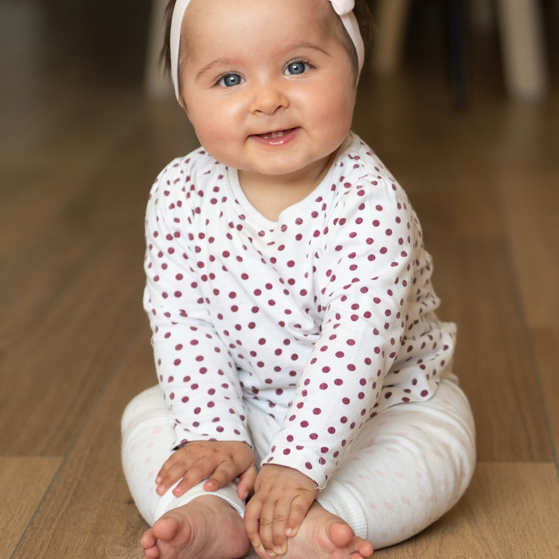 Smiling baby girl in a white and burgundy polka dot outfit sitting on a wood floor | Bundled gently-loved girl clothing bundles Canada, affordable one of a kind baby girl outfits from newborn to 24 months