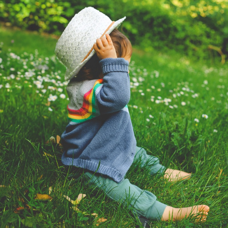 Toddler in a knit sweater trying on a sun hat in a daisy field | Bundled add-on accessories, gently-loved baby hats mitts and bibs in Canada
