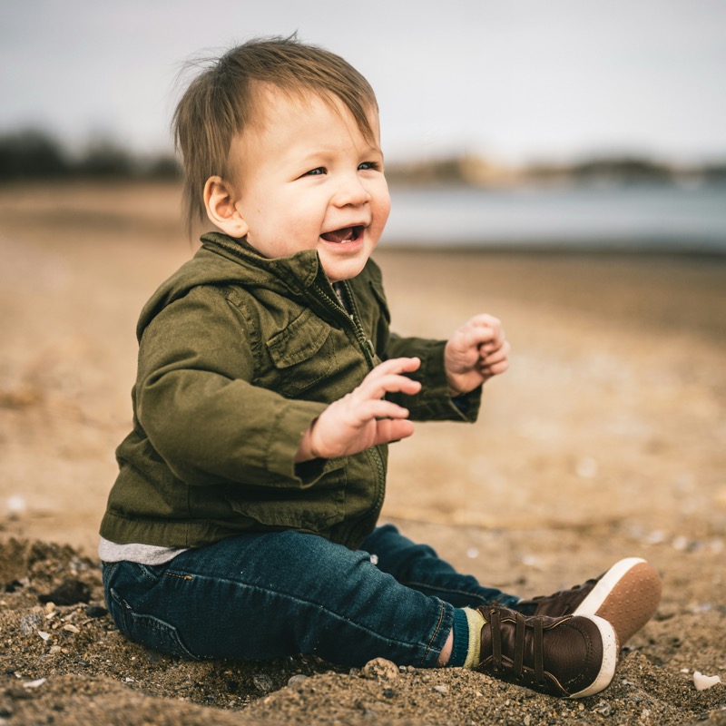 Laughing toddler in a green jacket sitting on a sandy beach | Bundled add-on outerwear, gently-loved baby and toddler jackets and coats in Canada